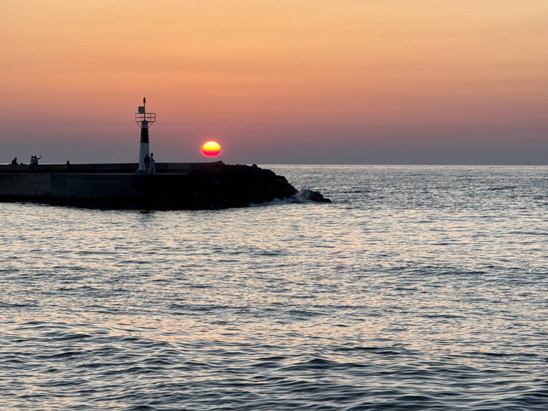 Auto-generated description: A serene sunset over the ocean with a lighthouse on a rocky pier.