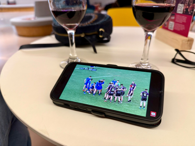 Auto-generated description: Two glasses of red wine are on a table next to a smartphone displaying a sports game.