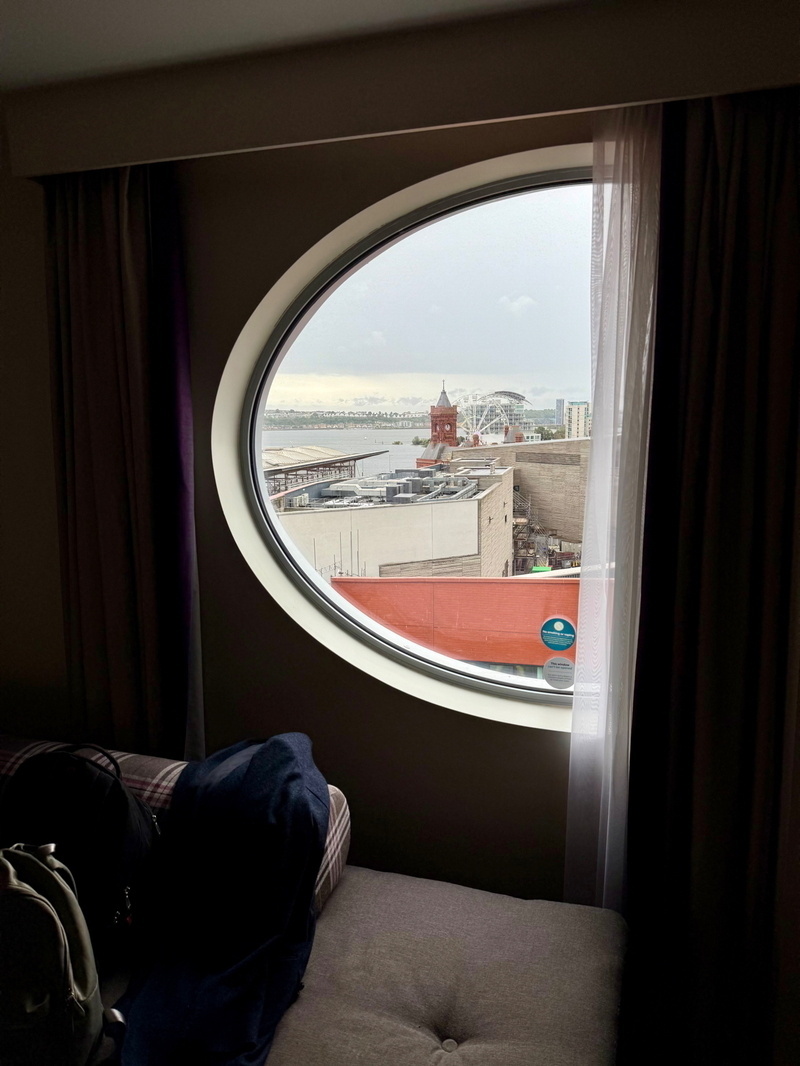 A circular window view from a room shows a waterfront scene with a red brick clock tower, nearby buildings, and a Ferris wheel against a cloudy sky. The foreground includes a cushioned bench with a backpack and a coat.