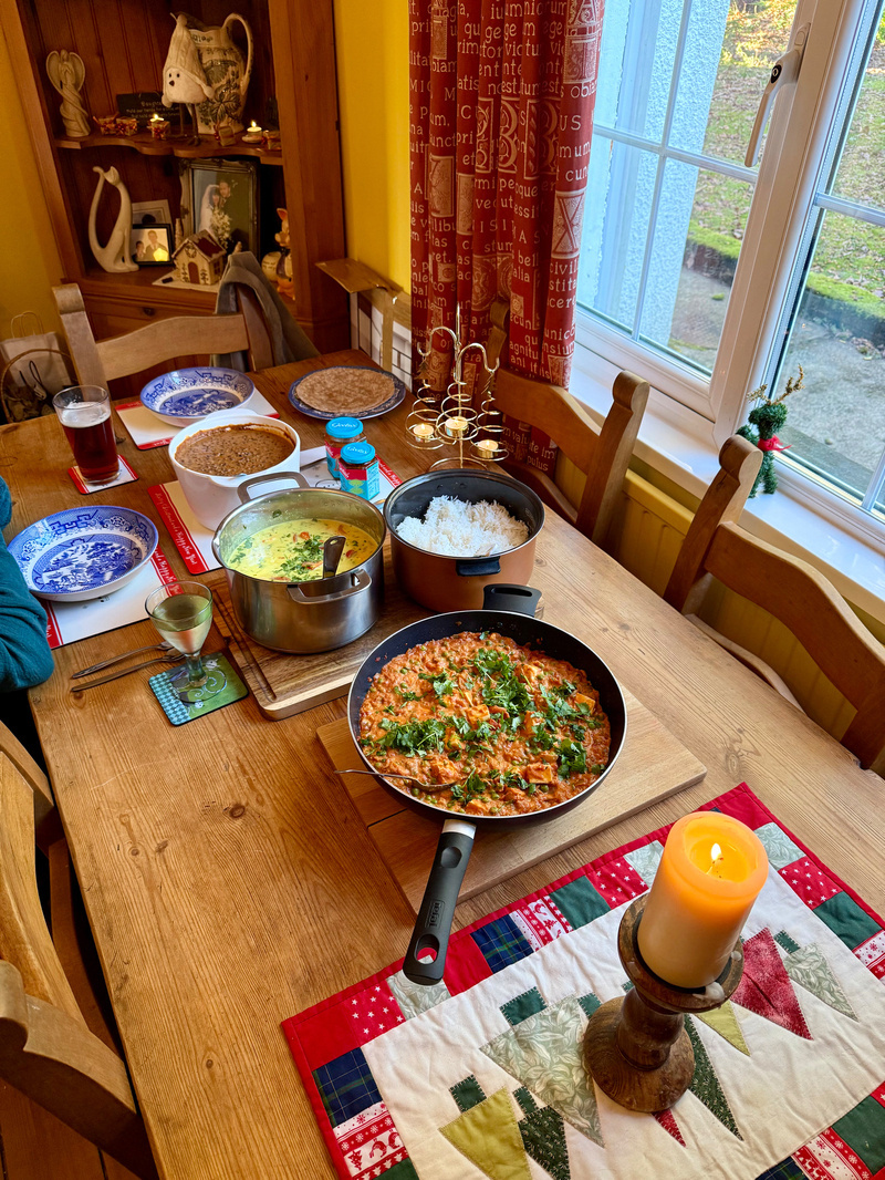 Auto-generated description: A cozy dining table is set with a variety of food dishes, including a pan of what appears to be paneer curry, alongside rice, bread, and drinks, with a decorative candle and a view of a garden outside the window.