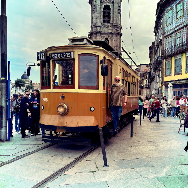 Auto-generated description: A vintage tram marked 18 Passeio Alegre is surrounded by people on a city street with historic buildings and a church tower in the background.