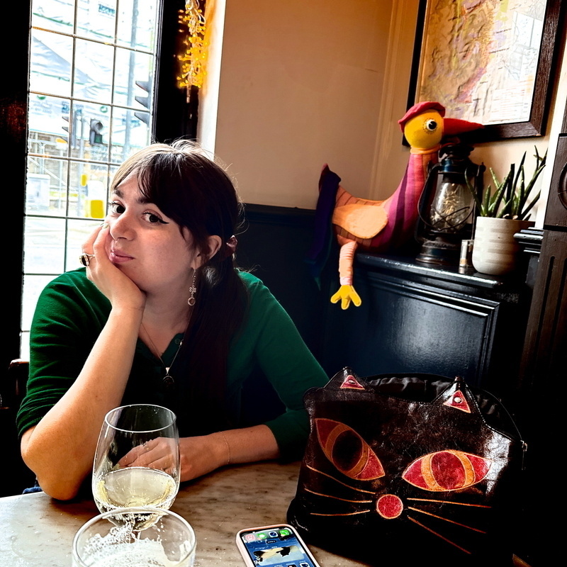 Auto-generated description: A woman in a green top sits at a table with two drinks, while a colorful bird sculpture decorates the background and a cat-faced purse rests nearby.