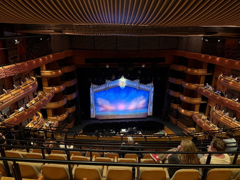 Auto-generated description: An ornate theater with a large stage features an intricate backdrop, while audience members are seated in the balconies and orchestra levels.