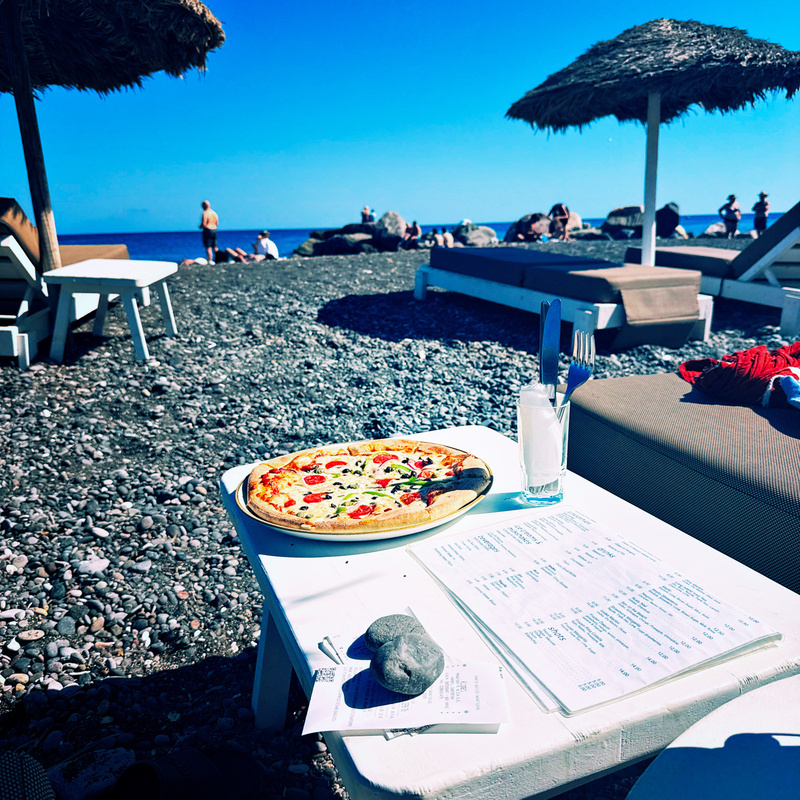 A pizza and a drink are set on a white table by a pebbled beach with sun loungers and umbrellas under a clear blue sky.