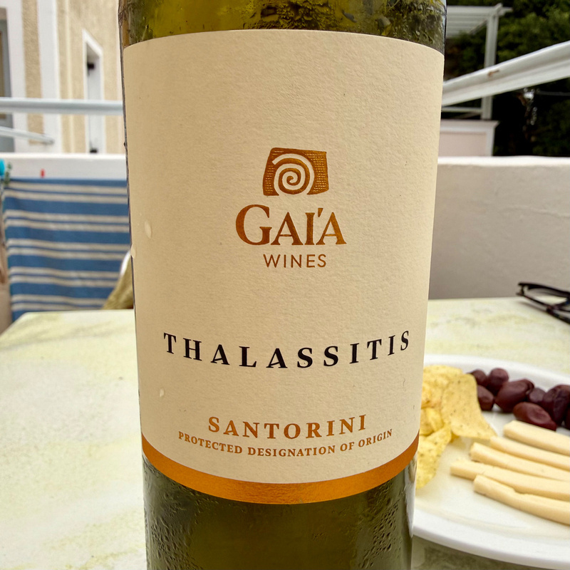 Auto-generated description: A bottle of Gaia Wines' Thalassitis from Santorini is displayed on a table alongside crisps, cheese, and olives.