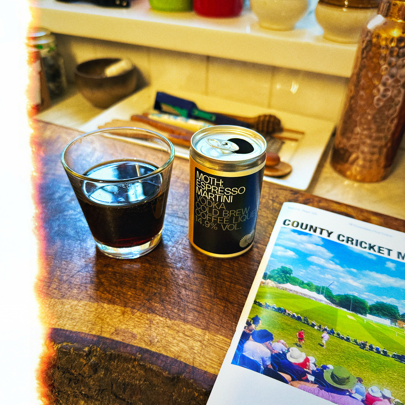A can of MOTH Espresso Martini and a glass of the drink sit on a wooden surface. Next to them is a magazine titled County Cricket Matters with an image of a cricket field on the cover. Various kitchen items are visible in the background
