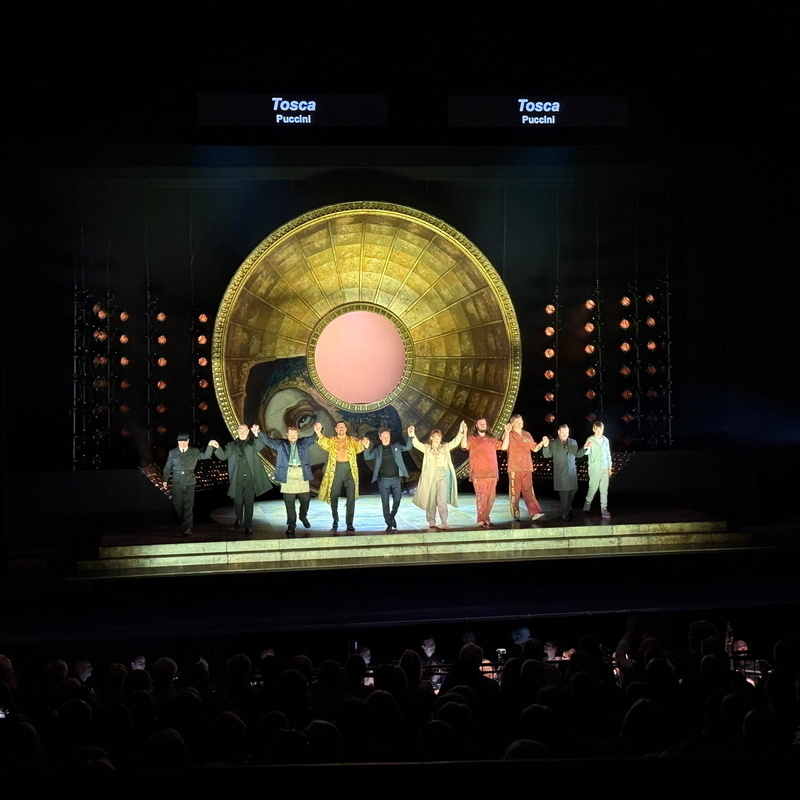 The image shows a group of performers taking a bow on stage after an opera performance of Puccini's Tosca.They stand in front of a large, circular, decorative backdrop with a central circular opening. Stage lights surround the backdrop.