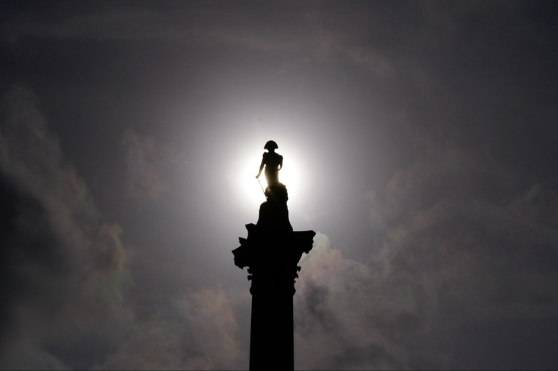 Auto-generated description: A silhouette of a statue stands atop a column against a dramatic sky with the sun creating a halo effect around it.
