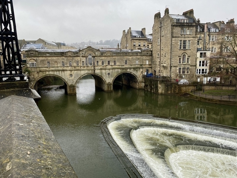 A historic stone bridge spans a river with cascading water, surrounded by old buildings in an overcast setting.