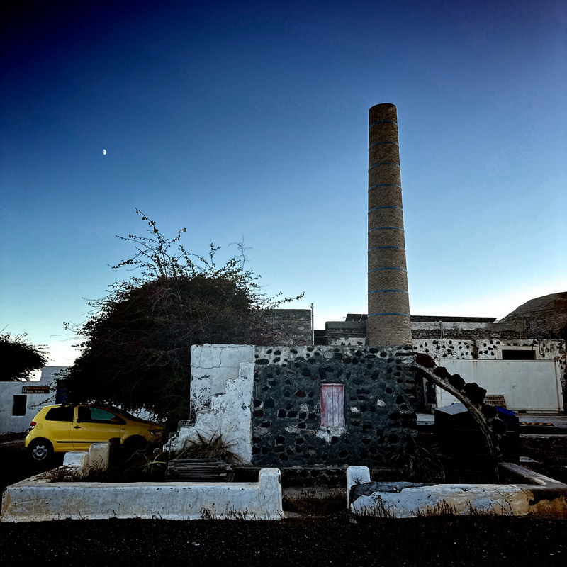 Auto-generated description: A rustic stone structure with a tall chimney stands beside a yellow car and a leafless tree under a darkening sky.
