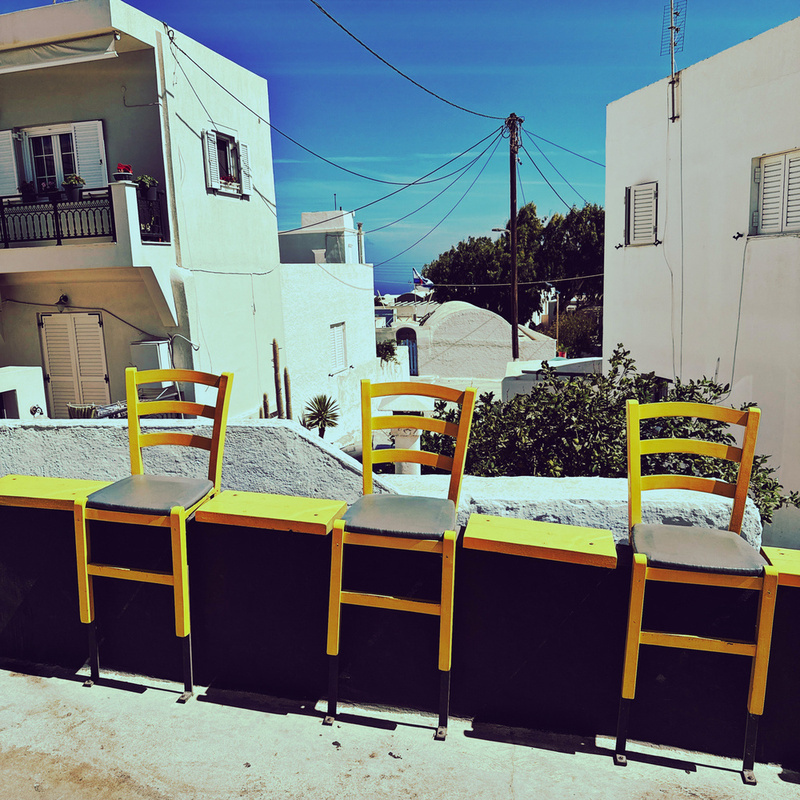 Three yellow chairs are lined up against a wall in a sunny street with white buildings and a clear blue sky.