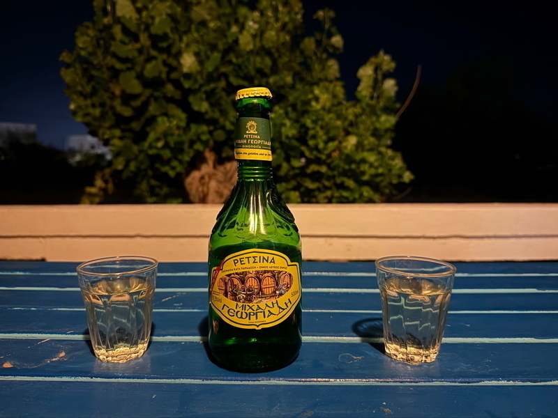 Auto-generated description: A bottle of Greek wine labeled Retsina is placed between two glasses on a blue table against a background of greenery at night.