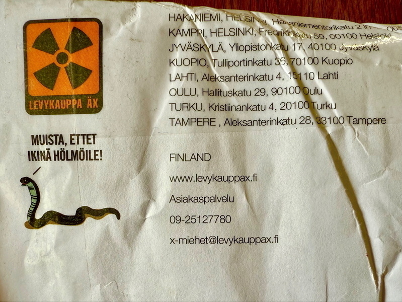 Auto-generated description: A wrinkled paper package features a radioactive symbol, several Finnish addresses, a cartoon snake, and text in Finnish alongside contact details for a business.