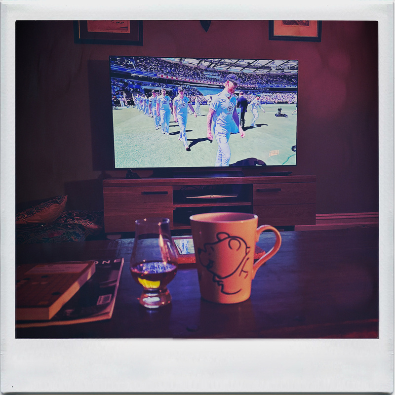 Auto-generated description: A living room scene features a television showing a cricket match, with a coffee table displaying a mug, a glass of whisky, and a book.