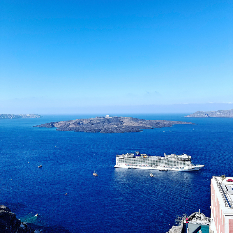 A cruise ship sails through a vast, clear blue sea with a hilly island in the background under a bright, cloudless sky.
