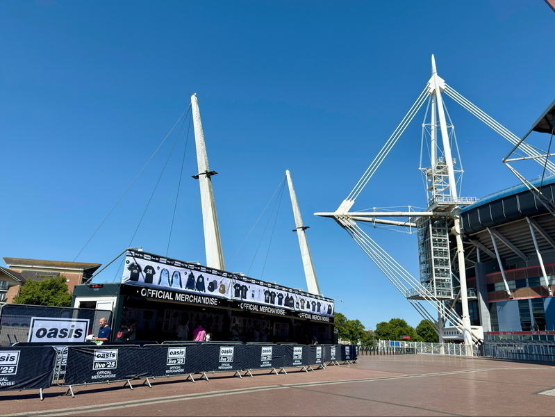 Auto-generated description: A merchandise stand for an event is set up outside a large sports stadium under a clear blue sky.