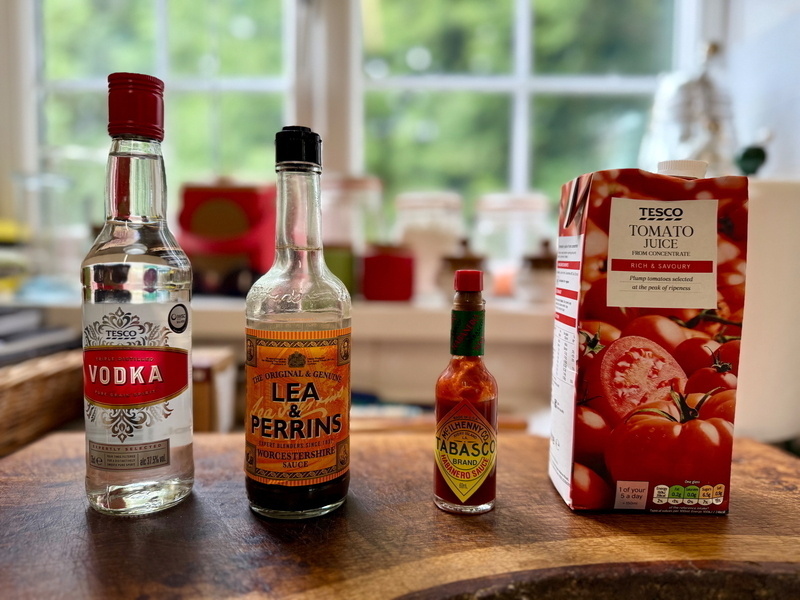 Auto-generated description: Bottles of vodka, Worcestershire sauce, Tabasco sauce, and a carton of tomato juice are lined up on a kitchen counter.
