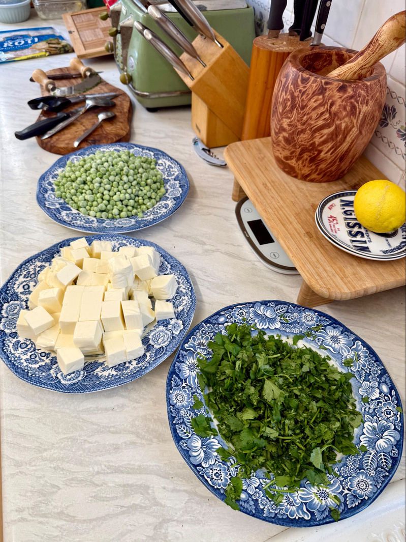 Auto-generated description: Various ingredients including peas, cubed paneer, and chopped coriander are arranged on blue floral plates in a kitchen setting.