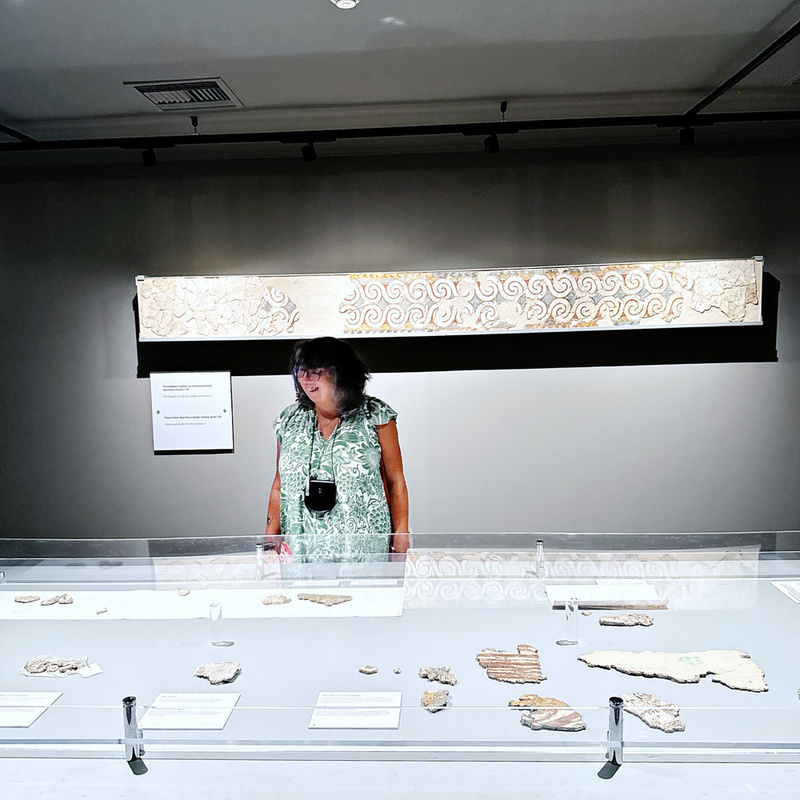 A person stands in a museum looking at ancient artifacts displayed in a glass case, with a long decorative stone piece mounted on the wall behind them.