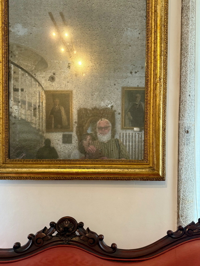 Auto-generated description: An ornate gold-framed mirror reflects a spiral staircase, chandeliers, paintings, and a person taking a photo in a room with a red upholstered chair.