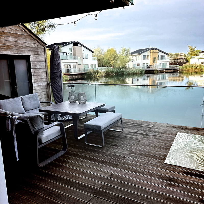 A modern lakeside deck with a wooden floor features a dining table set with cushioned chairs and benches. Two lanterns sit on the table. The deck overlooks a peaceful lake surrounded by contemporary houses with large windows.
