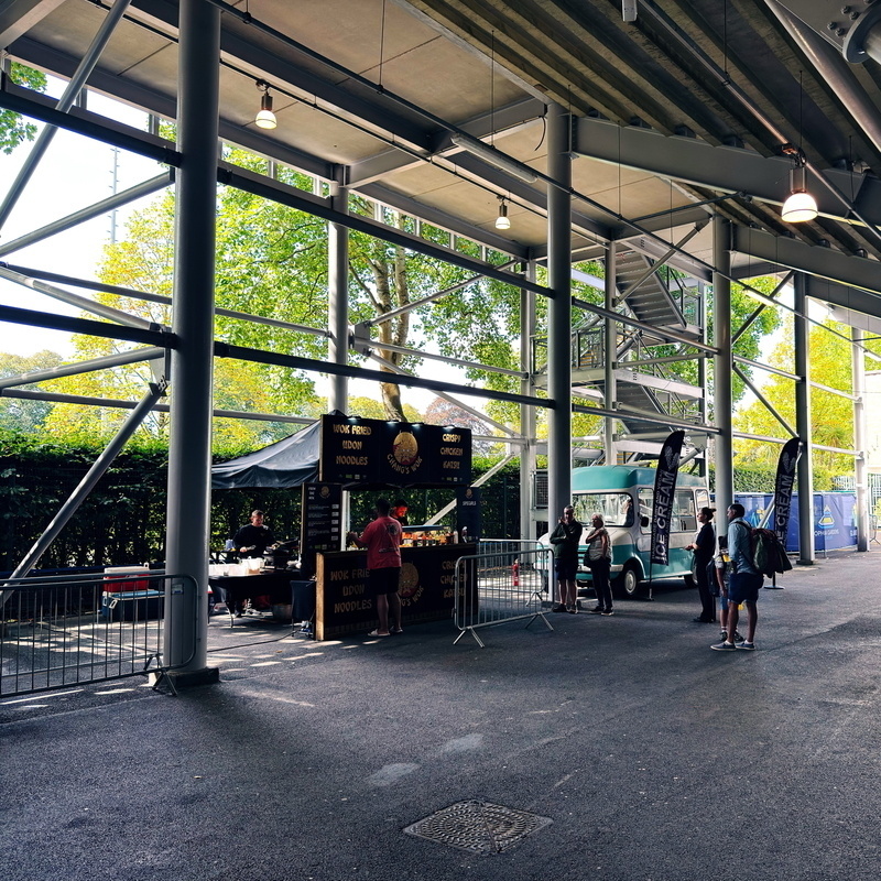 A food stall under a structure with large windows and metal framework. The stall offers wok-fried udon noodles and katsu. People are lined up at the counter, and a vintage ice cream van is parked nearby.