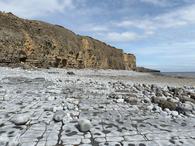 Auto-generated description: A rocky coastline with large cliffs and scattered stones under a partly cloudy sky.