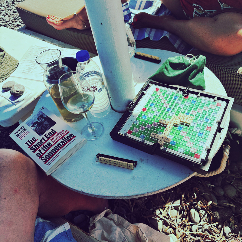 A relaxing outdoor scene depicts a Scrabble board game, books, a glass of wine, and people lounging on a stony beach.