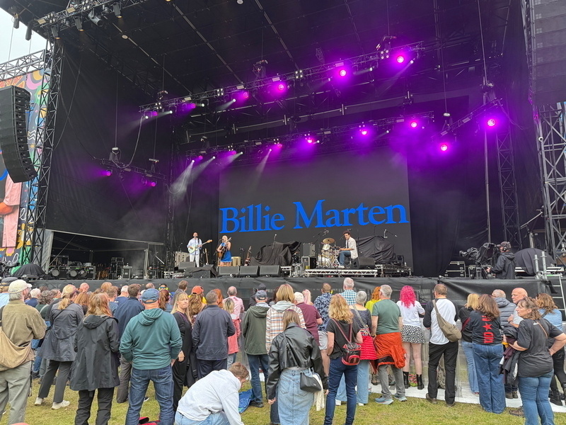 Auto-generated description: A band performs on an outdoor stage with a sign reading Billie Marten, while a crowd watches under purple stage lights.
