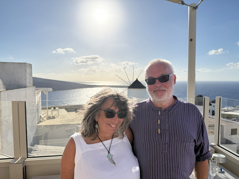 Auto-generated description: A smiling couple wearing sunglasses poses in a sunny seaside setting with a windmill and ocean in the background.