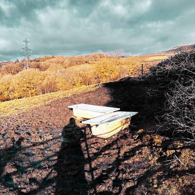 A wheelbarrow sits on a muddy hill overlooking a landscape with leafless trees and a cloudy sky.