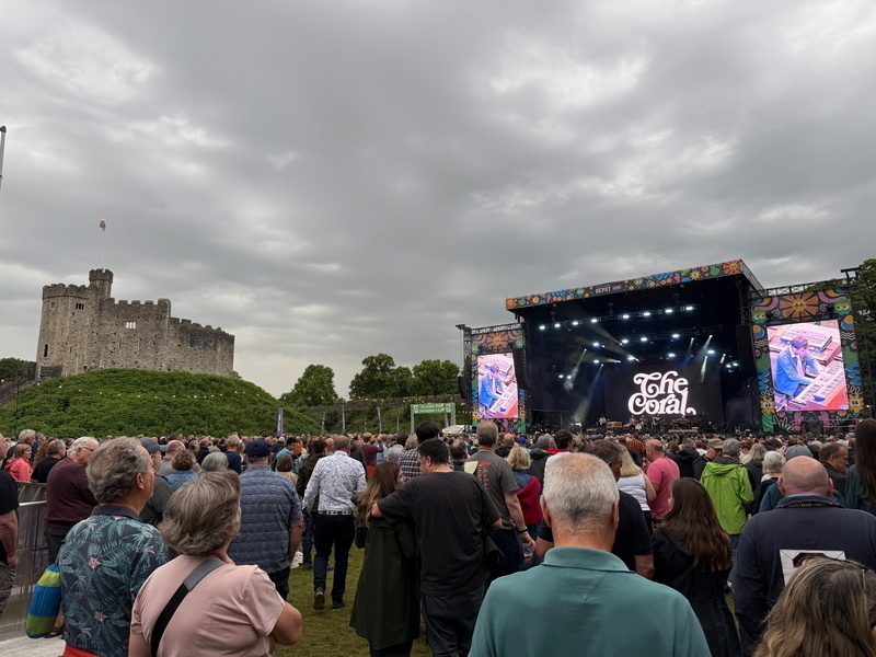 Auto-generated description: A large crowd gathers at an outdoor concert near a historic castle under a cloudy sky.