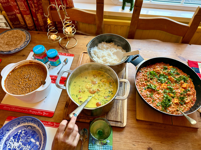 Auto-generated description: A table is set with several dishes, including a pot of soup or stew, rice, a pan of curry or vegetable dish, and sides, with a hand reaching for one of the pots.