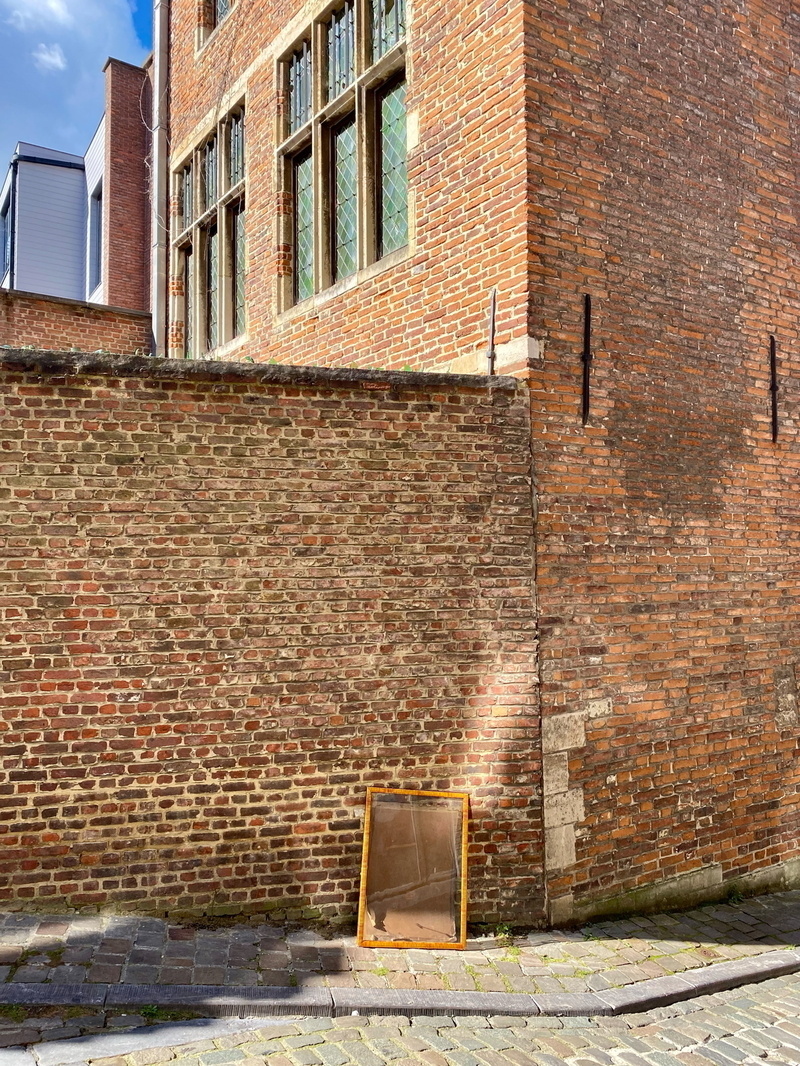 Auto-generated description: A large brick building with a tall wall has a framed mirror propped against it on a cobblestone street.
