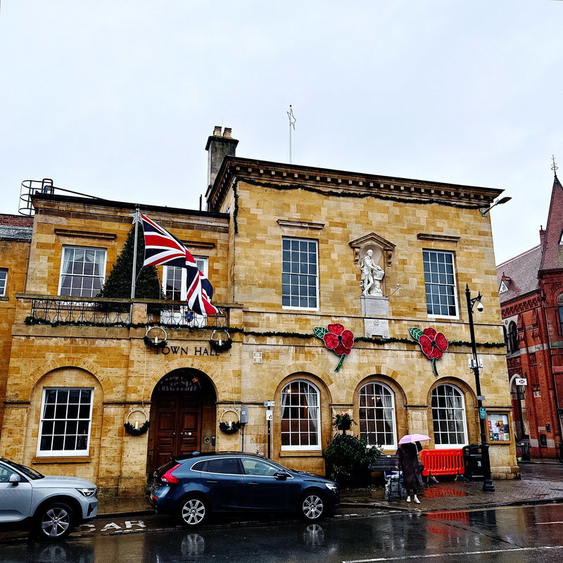 Auto-generated description: A classic stone town hall adorned with a large Union Jack flag and remembrance poppy decorations is set against a rainy street with passing pedestrians and parked cars.