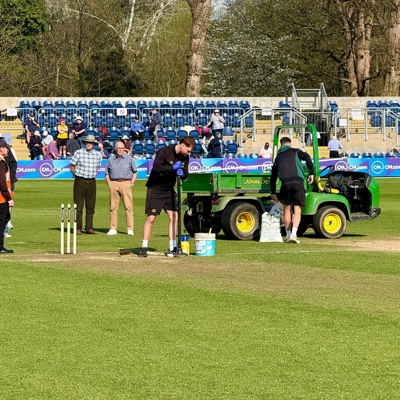 Auto-generated description: Several people are preparing or maintaining a sports field, with one person in front of cricket stumps and a tractor nearby.