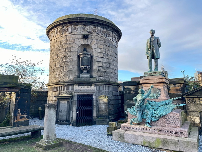 Auto-generated description: A historical stone monument and a statue dedicated to Scottish-American soldiers are set against a partly cloudy sky.
