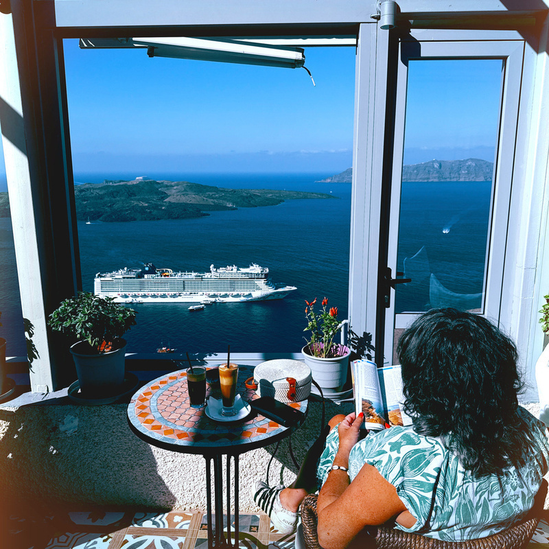 A person is relaxing with a book and drinks on a balcony overlooking the sea and a cruise ship.