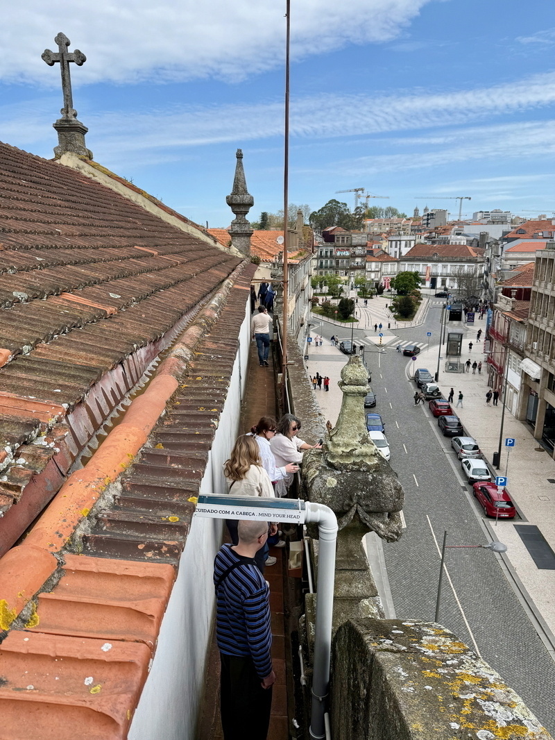 Auto-generated description: People are walking along a narrow ledge on a rooftop next to a street with parked cars and distant city views.