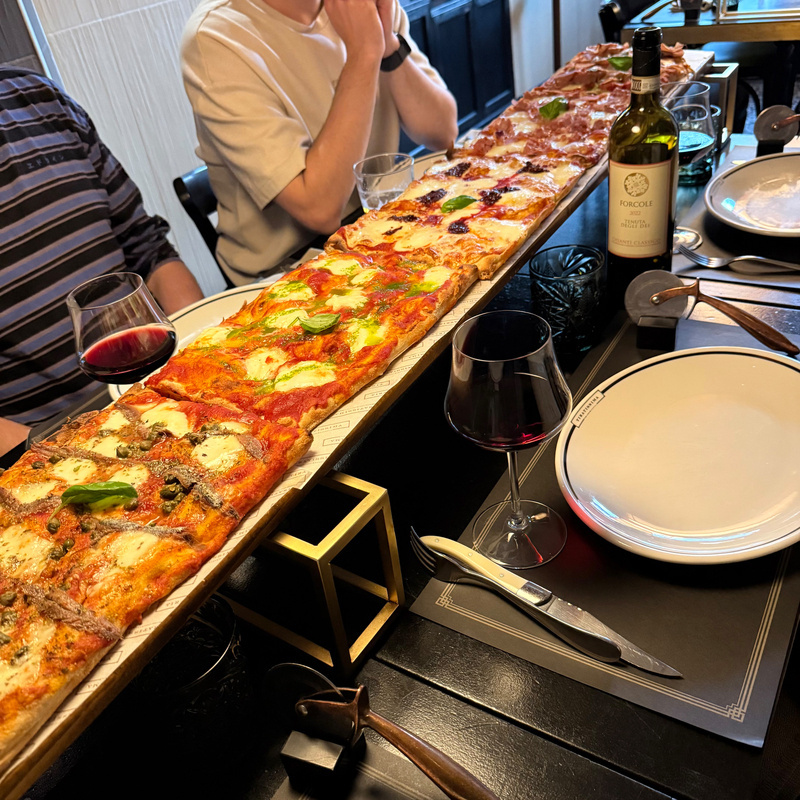 A long wooden board holds a variety of pizzas with different toppings, surrounded by wine glasses and plates set on a dining table.