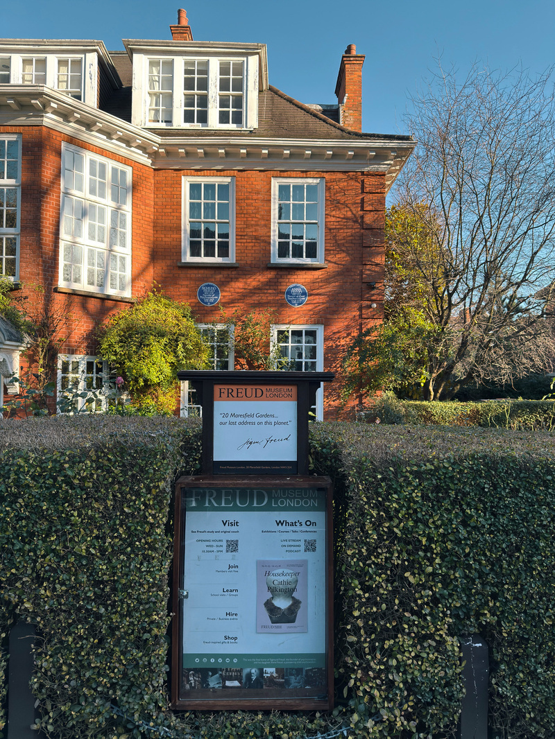 A red-brick building with white-framed windows is surrounded by trees and greenery, with a signboard for the Freud Museum at the forefront.