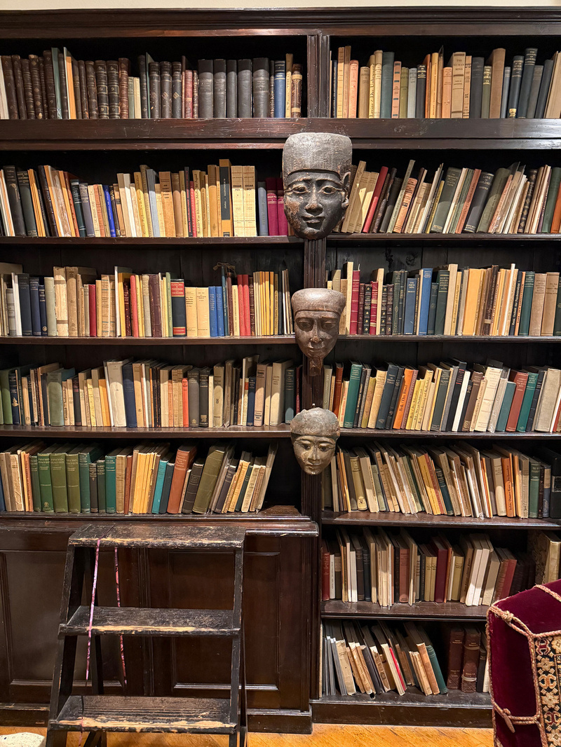 A bookshelf filled with various books is adorned with three sculpted faces vertically aligned in the center, accompanied by a wooden step ladder in the foreground.