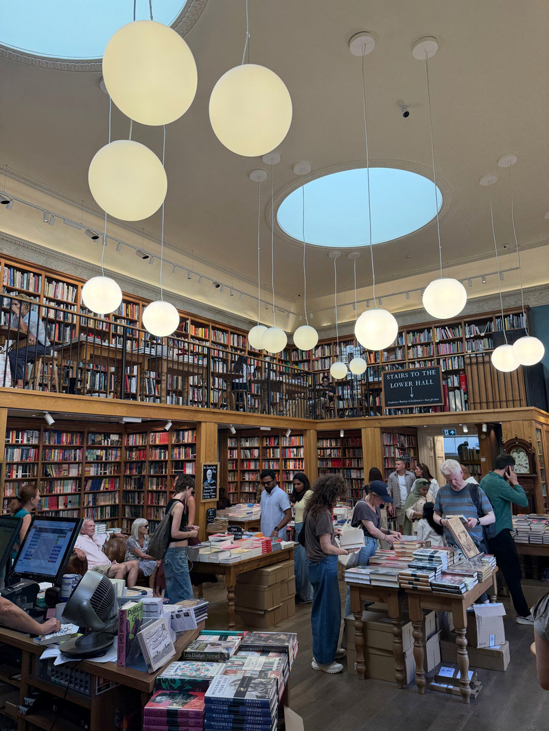 A spacious and brightly lit bookstore filled with shelves of books, tables stacked with more books, and customers browsing and engaging with the store.