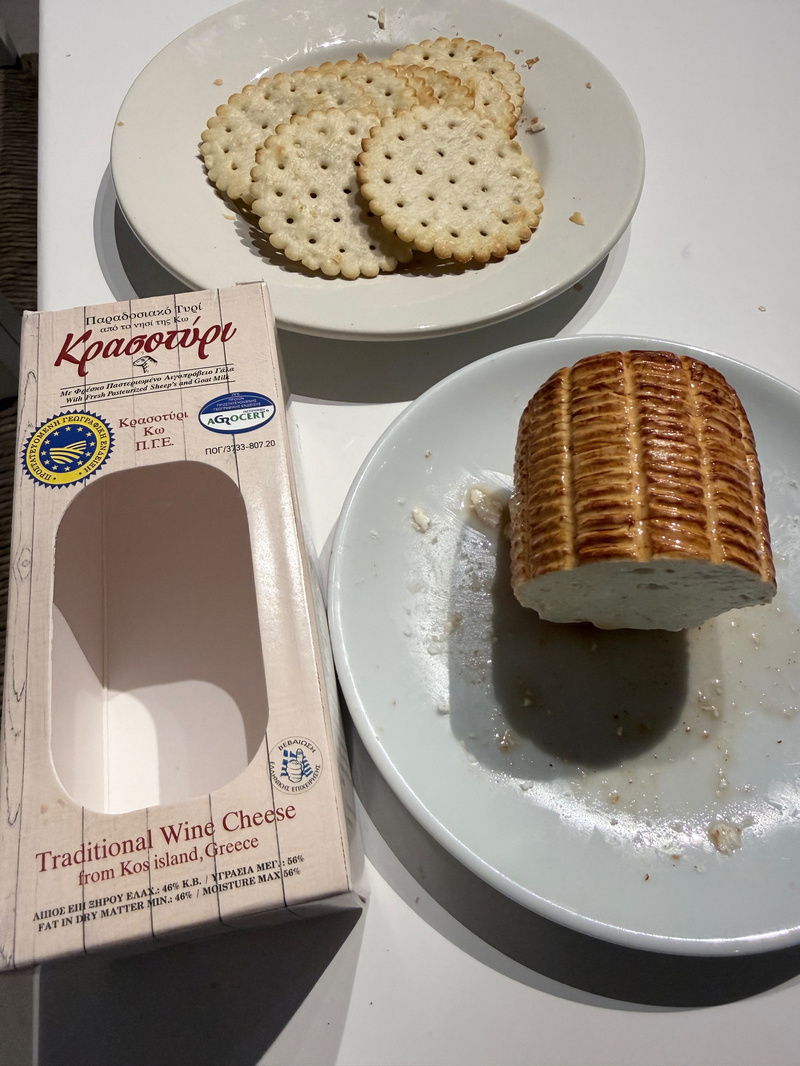 A plate of round crackers, a partially eaten piece of traditional wine cheese from Kos island, Greece, and its empty packaging are displayed on a table.