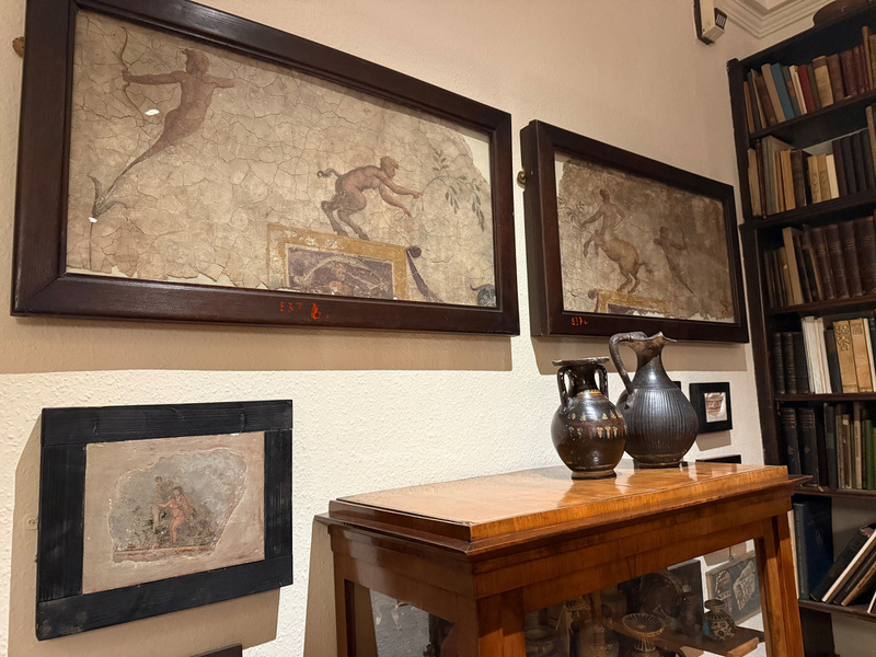 A room features framed ancient art on the walls, clay vessels on a wooden cabinet, and a bookshelf filled with books.
