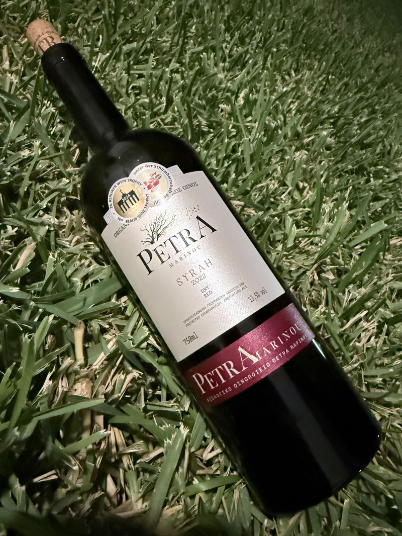 A bottle of Petra Marinas Syrah 2022 red wine is lying on green grass.