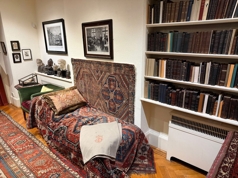 A room featuring a couch draped with patterned textiles, surrounded by bookshelves, framed pictures, and busts on a shelf.