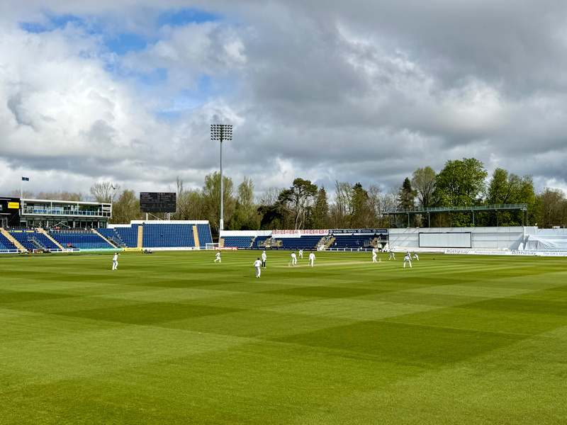 A cricket match is taking place on a green field under cloudy skies, surrounded by stadium seating.