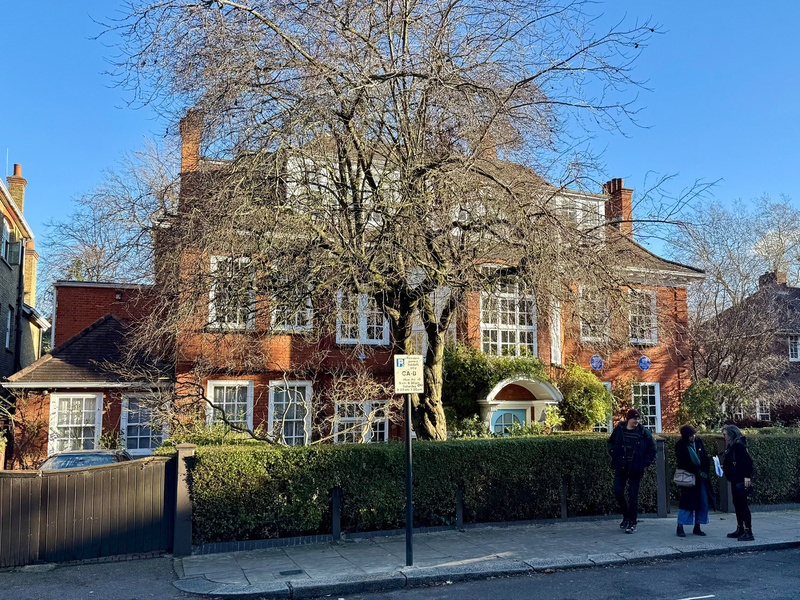 A large red brick house with multiple windows is partially obscured by a bare tree, while three people stand on the sidewalk in front of a hedge and a sign.