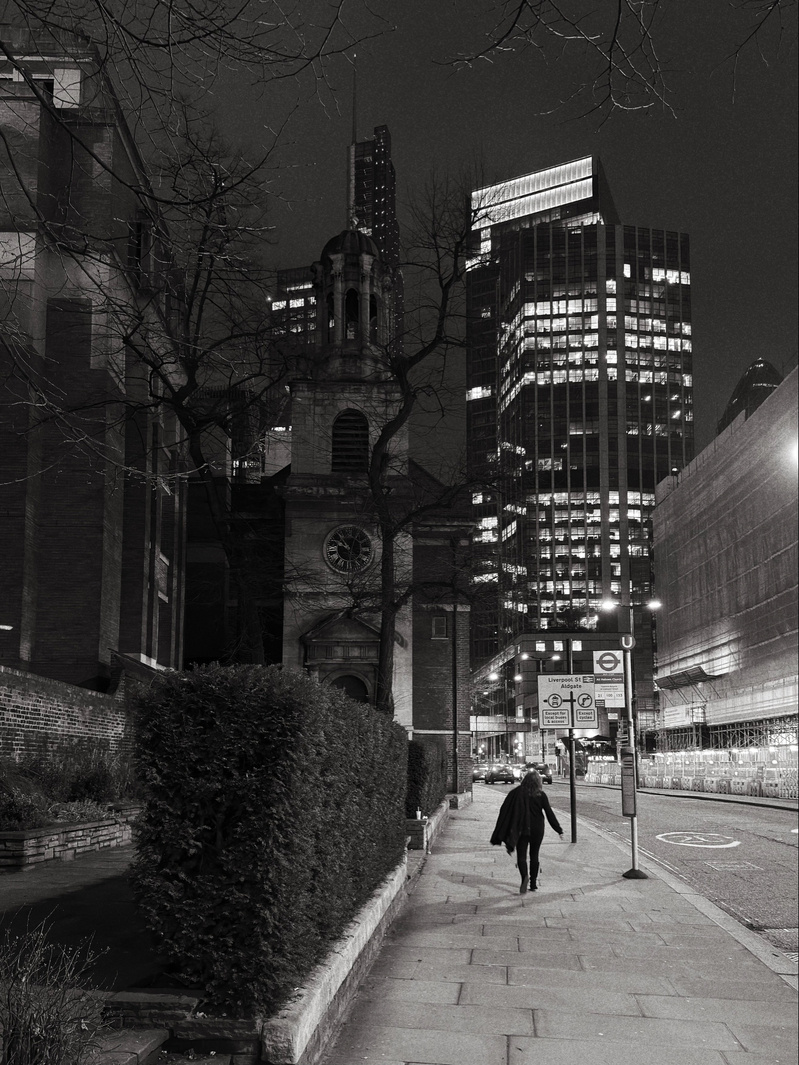 Auto-generated description: A solitary figure walks along a city street at night, framed by towering buildings and an old church.